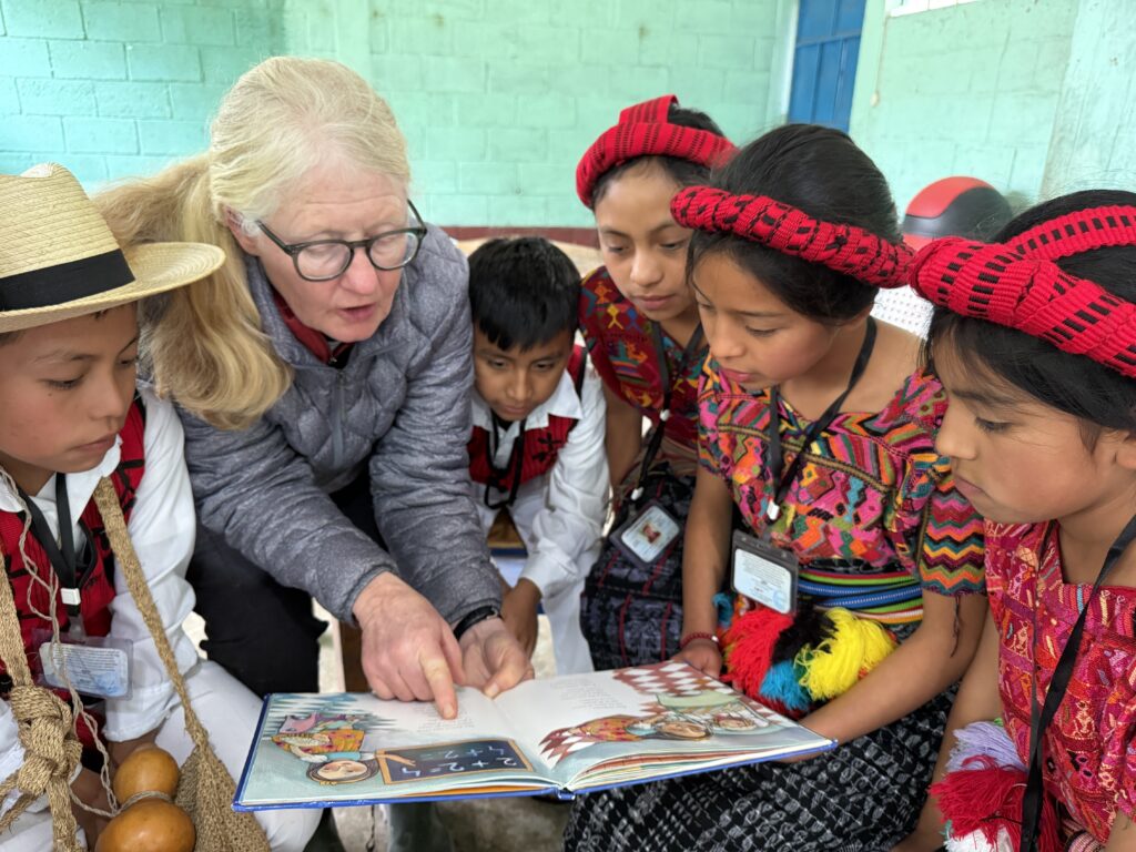 "Mission team member reading a book to a group of boys and girls at a village school. The children are attentively listening, dressed in vibrant traditional clothing, creating a heartwarming scene of cultural connection and education."